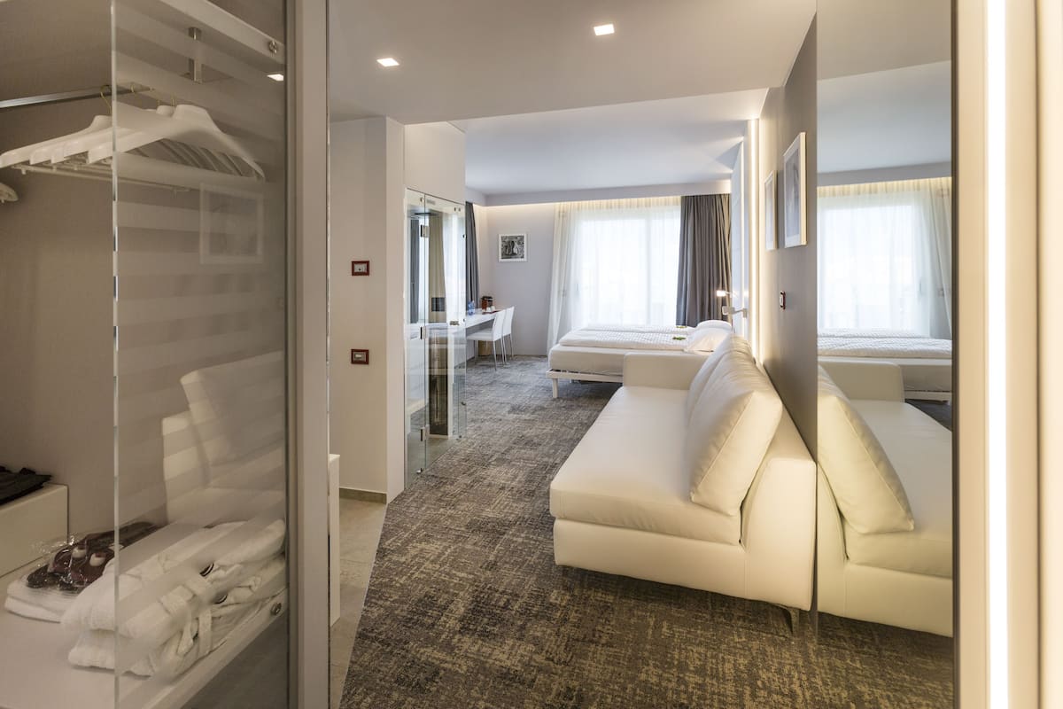 A modern hotel room with a glass-enclosed closet, a white sofa, a desk with a chair, and a bed with a white headboard.
