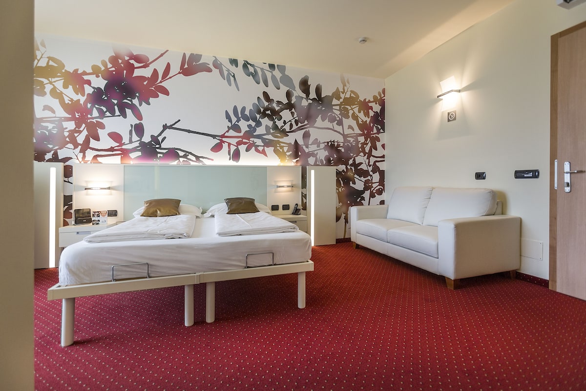 A hotel room with a bed, a white sofa, a red carpet, and a wall with a leaf pattern.