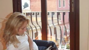 A person sitting on a bed with a colorful bedspread, reading a book, with a view of buildings through a window. - Mamma Maria (Taormina)