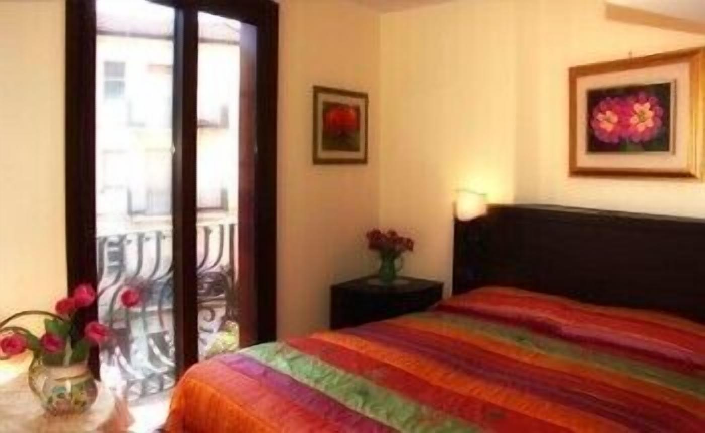 Una habitación de hotel con cama, mesita de noche, un jarrón con flores y ventana con vista a edificios.
