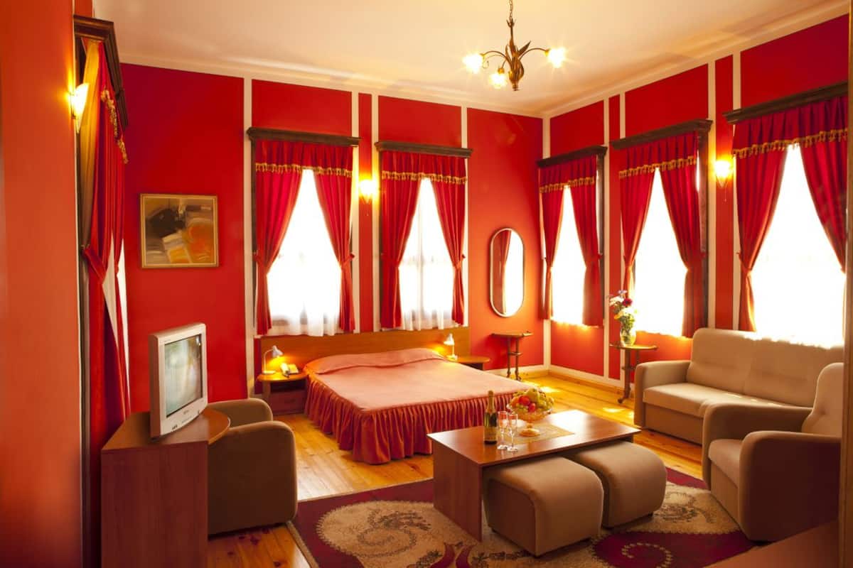 A hotel room with a red theme, featuring a bed, a sofa, a TV, and a dining table.