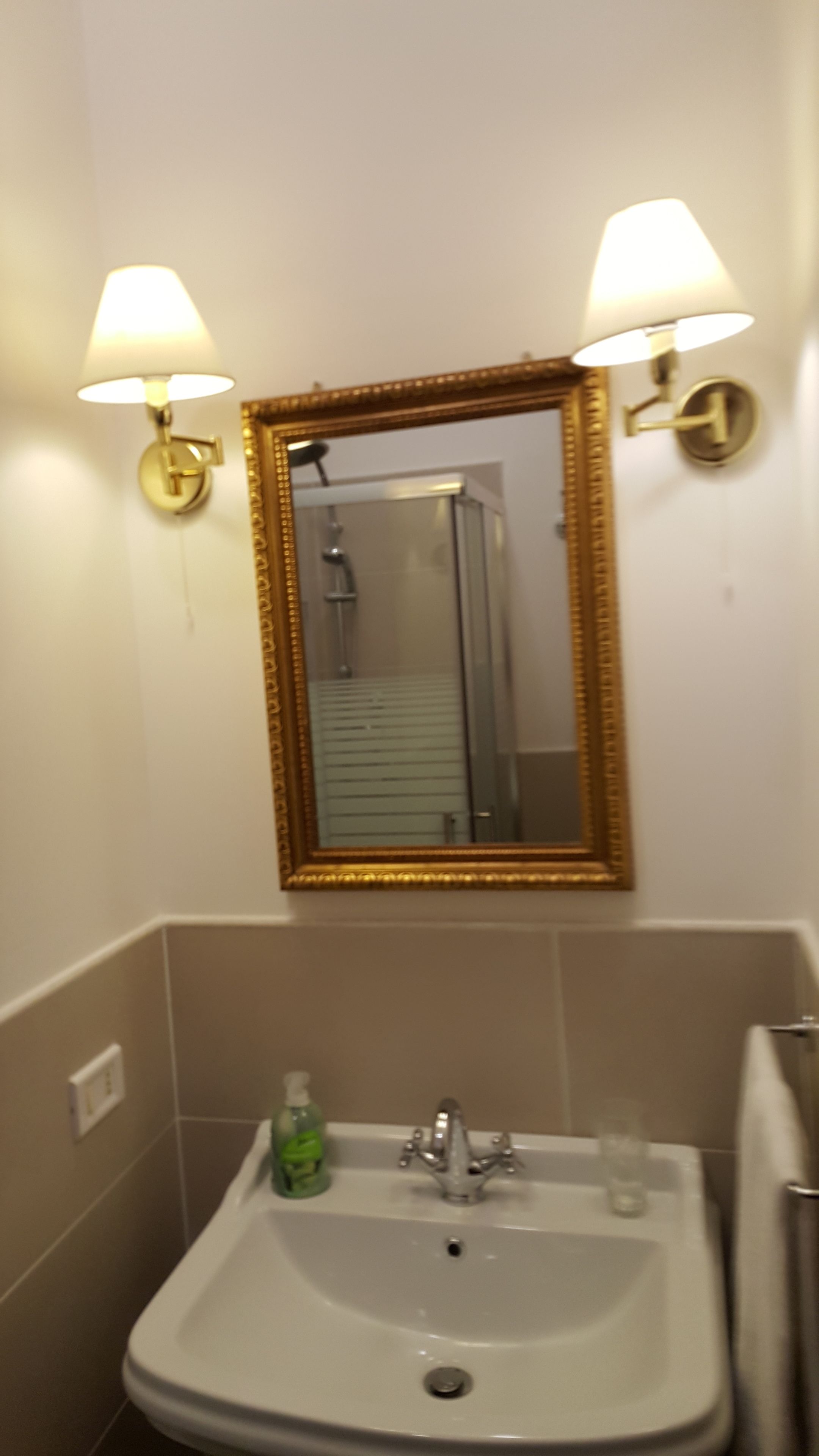 Un baño con un lavamanos blanco, dos lámparas fijadas a la pared y un espejo grande con marco dorado.