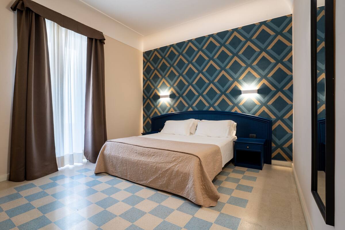 A hotel room with a checkered floor, a bed with a beige bedspread, a blue headboard, and a patterned wall.