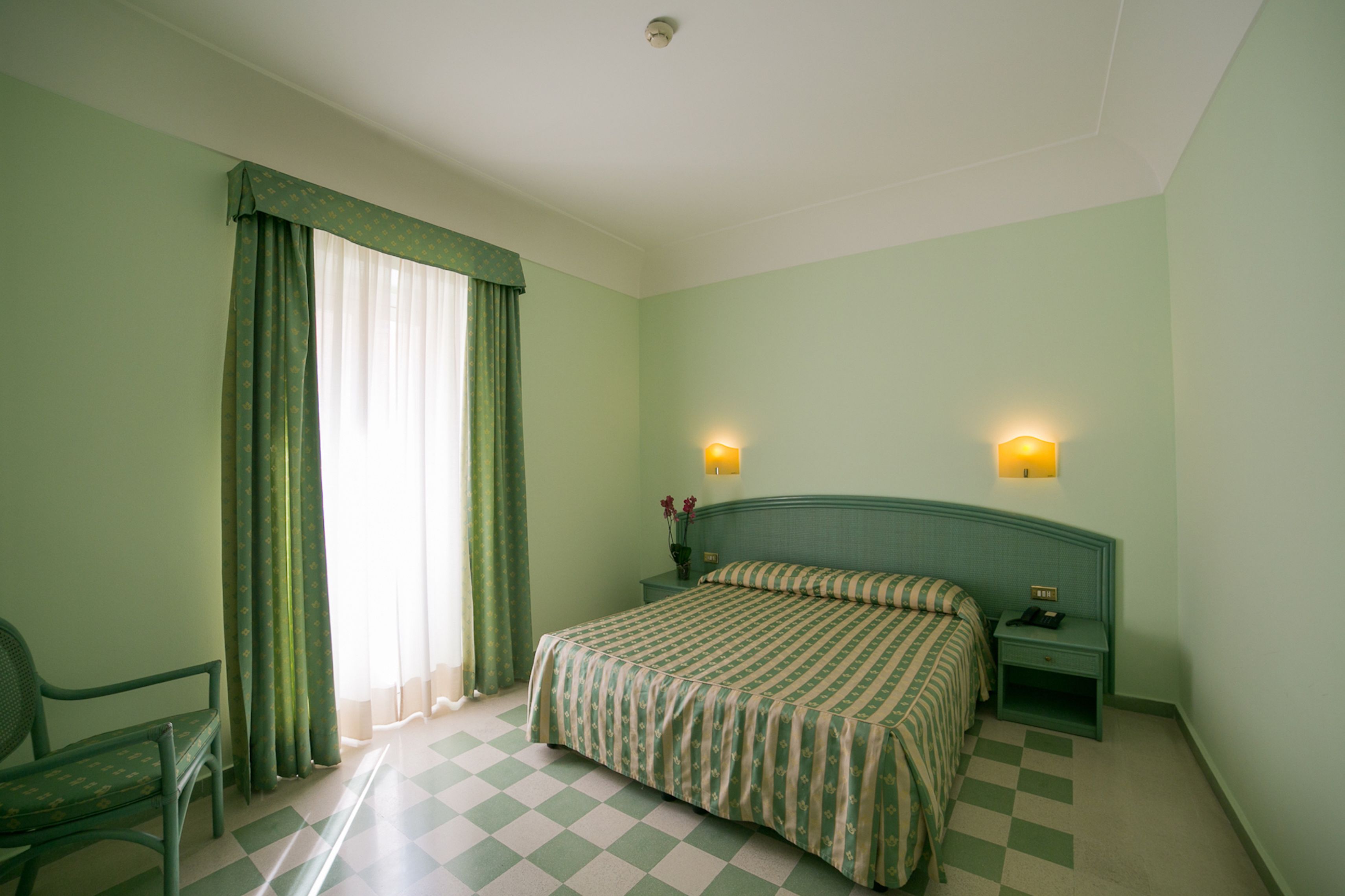 Ein Zimmer mit Schachbrettmuster-Boden, einem Bett mit gestreifter Bettwäsche, einem Sessel, einem Nachttisch mit Lampe und einem Fenster mit Vorhängen.