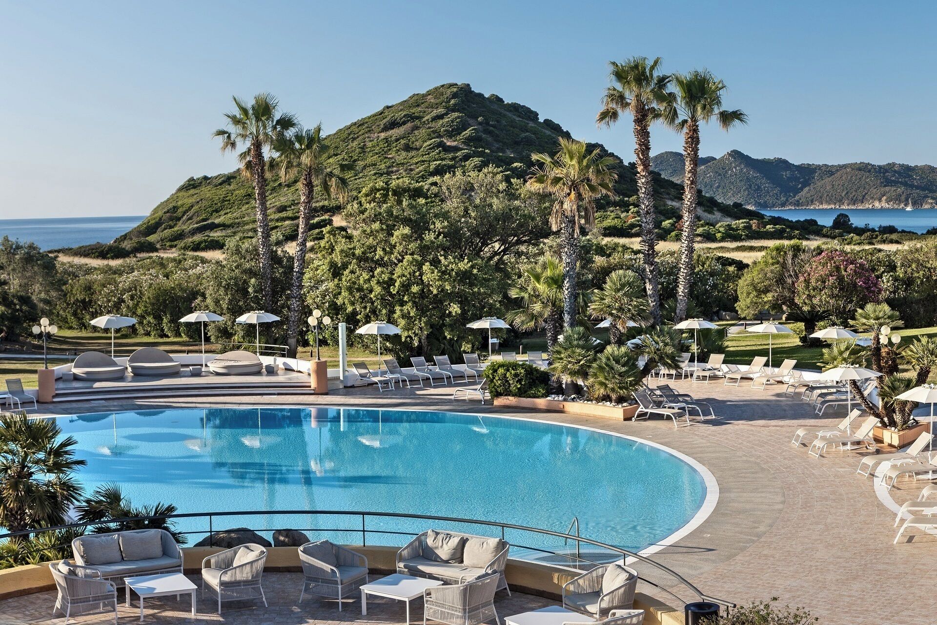 A pool area with lounge chairs and umbrellas, surrounded by palm trees and a view of the sea.