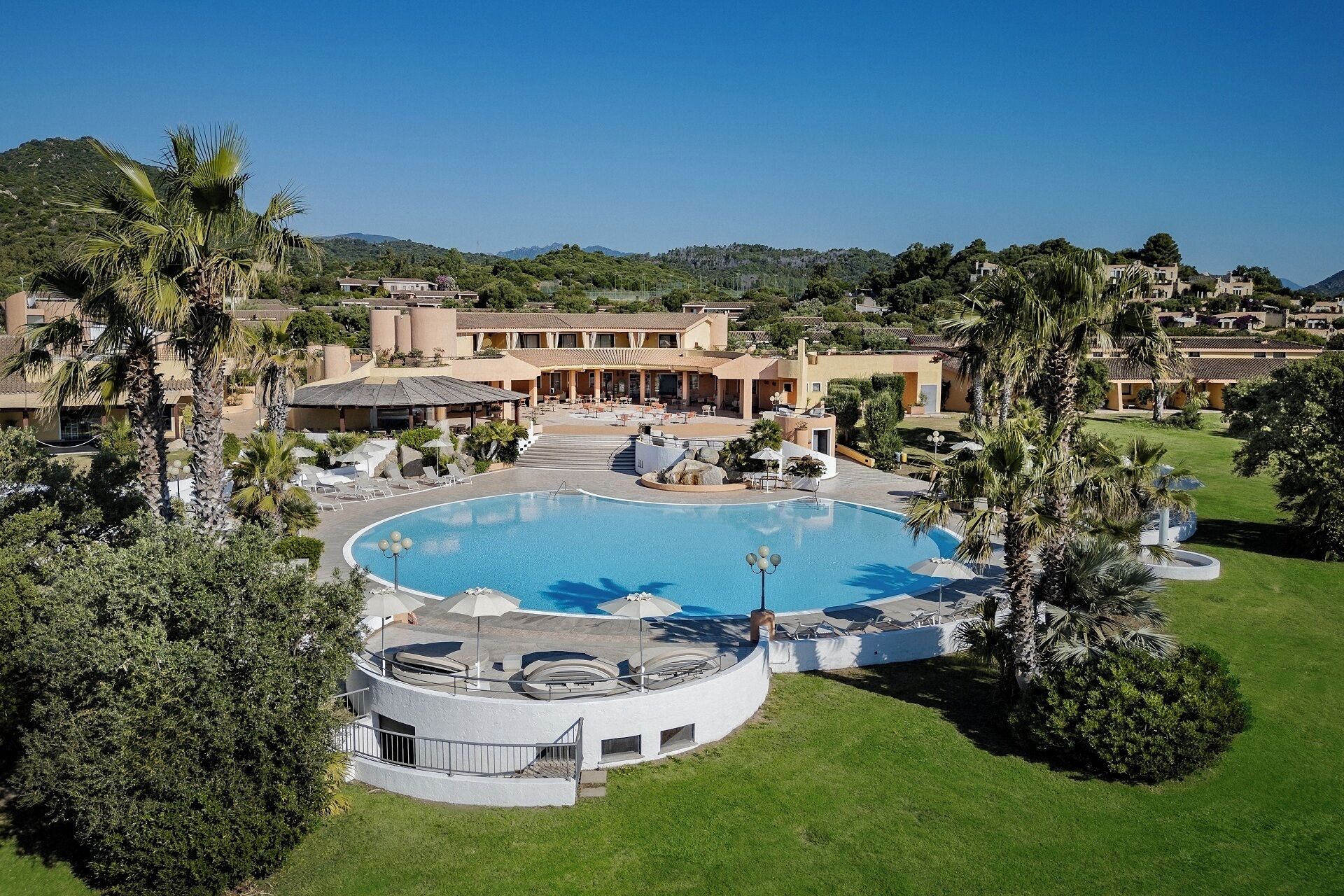 Un resort con piscina central, palmeras y montañas al fondo.