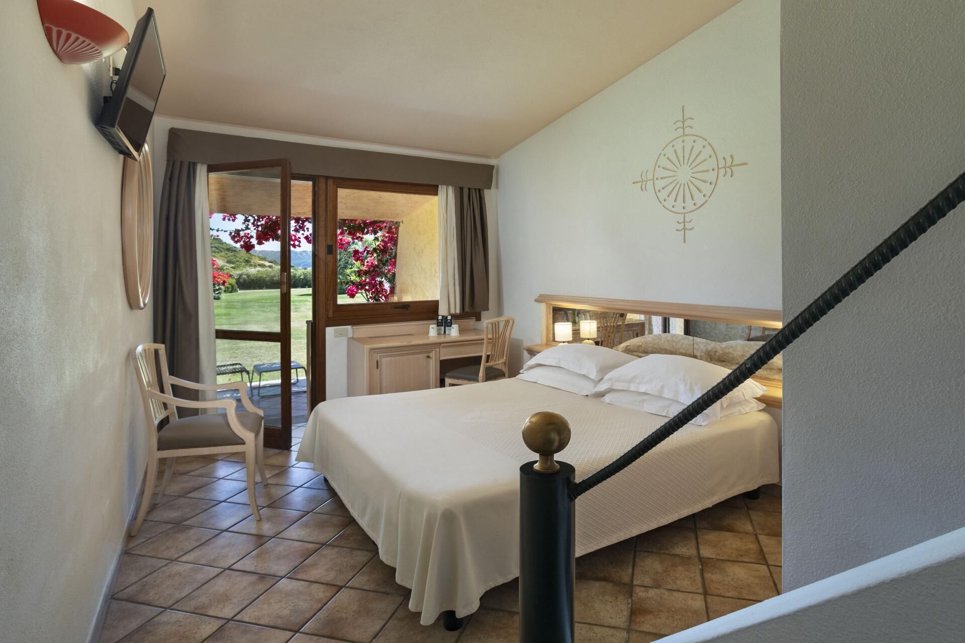 Camera d'albergo con un letto, una scrivania, una sedia e vista su un giardino attraverso una porta vetrata.