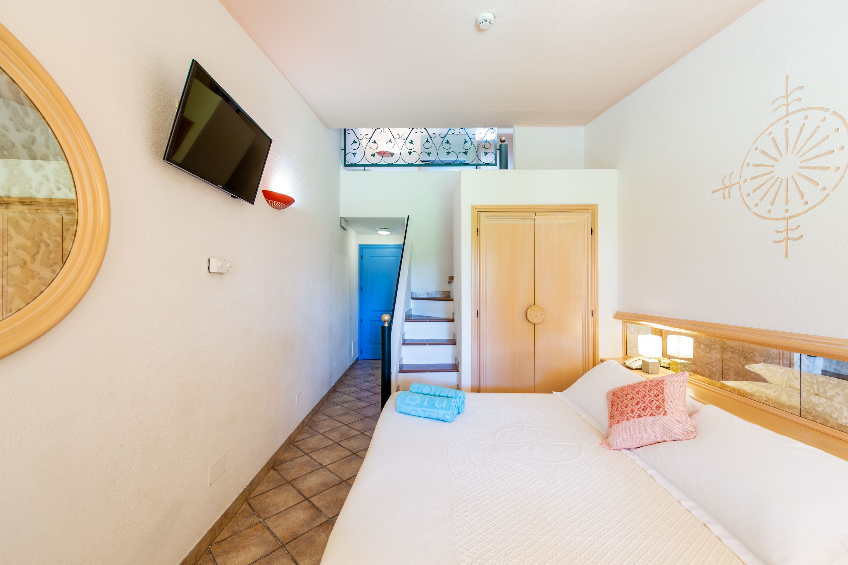 Une chambre à coucher avec un lit, un téléviseur fixé au mur, un escalier menant à une autre pièce et une porte.