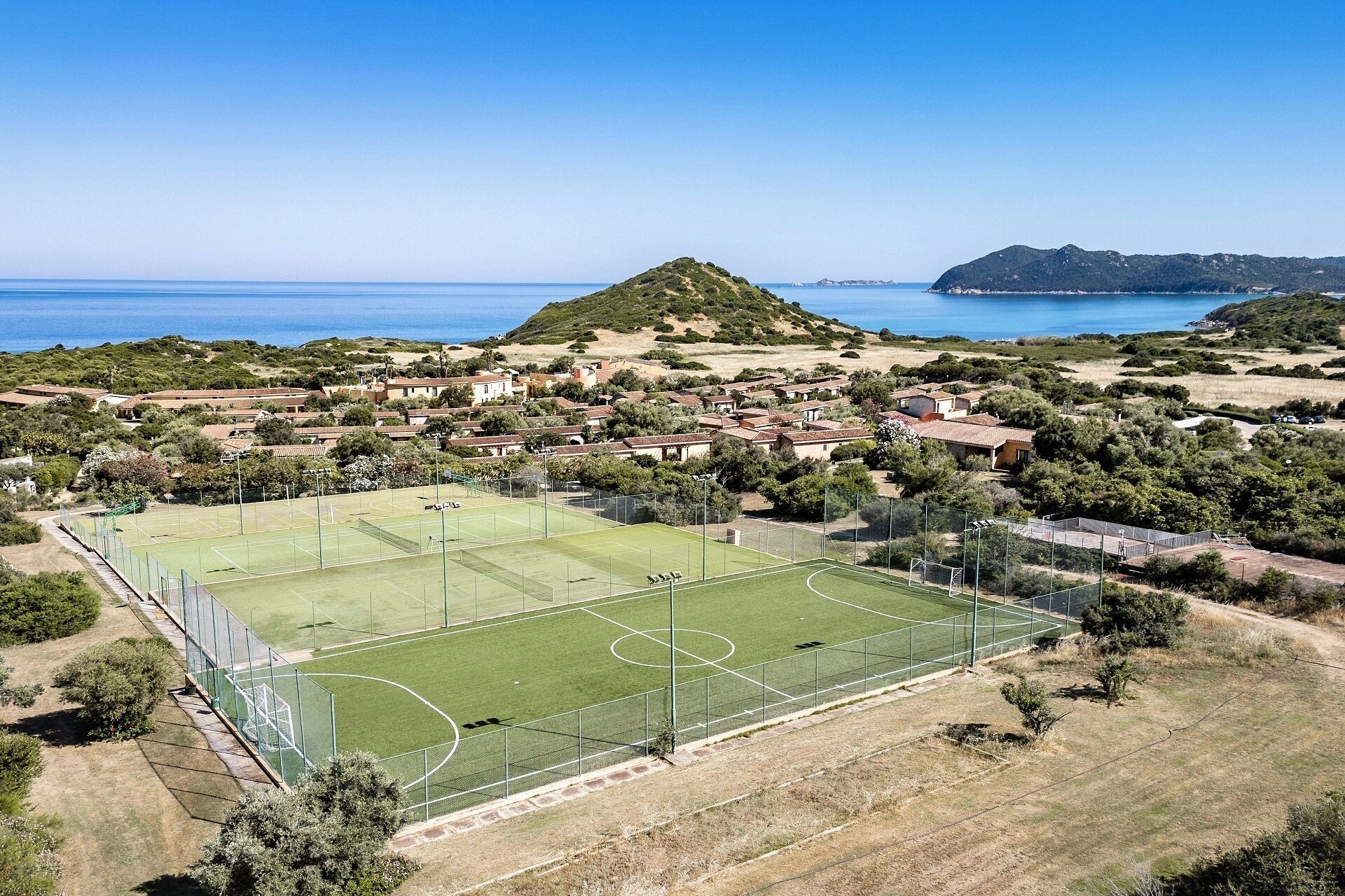 Vue aérienne d’un quartier résidentiel situé sur la côte, avec un court de tennis entouré de maisons et de verdure.