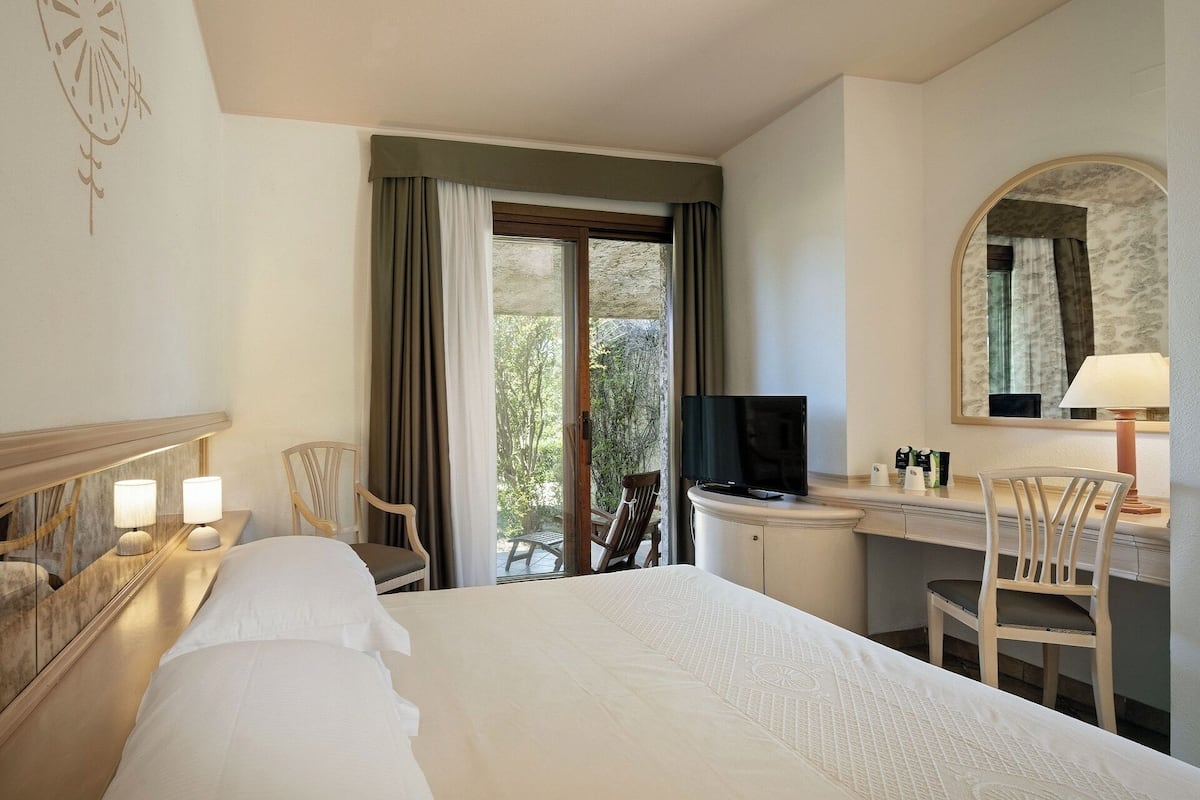 A hotel room with a bed, a desk with a TV, a chair, and a window with curtains.