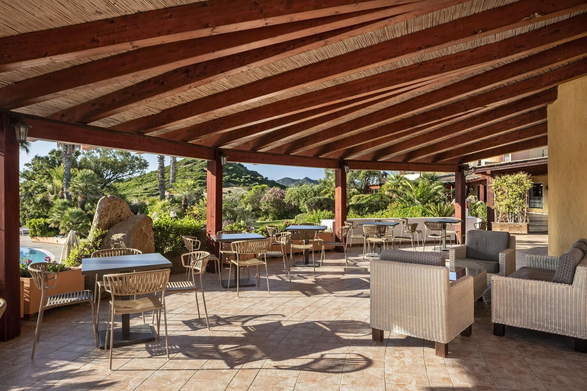 An outdoor patio with wooden beams, wicker furniture, and a view of a mountainous landscape.