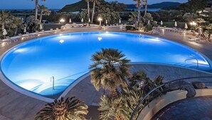 A large, curved swimming pool surrounded by palm trees and lounge chairs, with a view of the sea and hills in the background. - Sant'Elmo Beach Hotel (Castiadas)