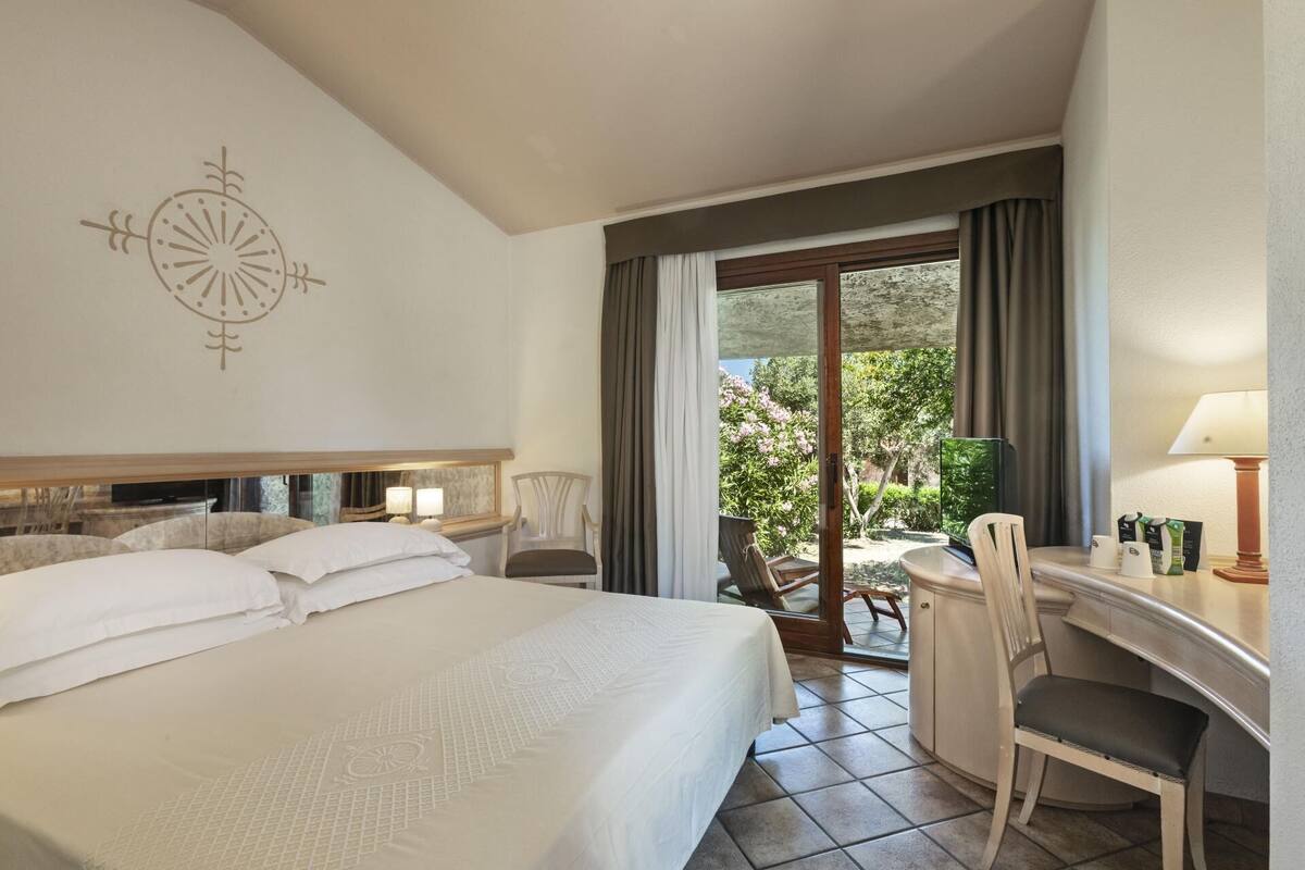 A hotel room with a bed, a desk, a chair, a lamp, and a view of the outdoors.