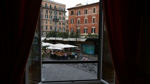 Vista desde una ventana con cortinas rojas, que da a una calle con terrazas al aire libre y un edificio de varios pisos con balcones.