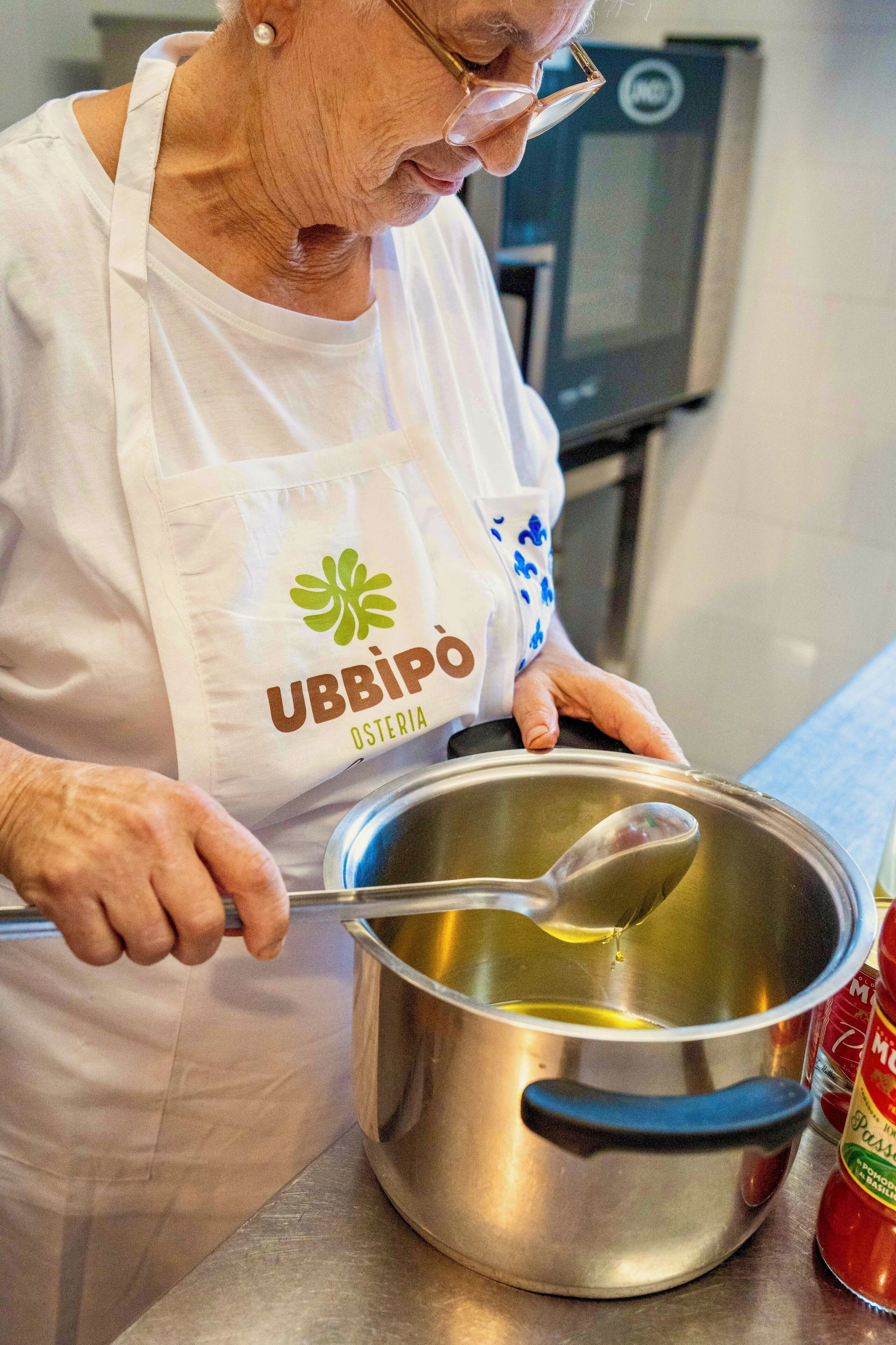 Una persona mayor con delantal blanco y un logo, removiendo una olla sobre la estufa.