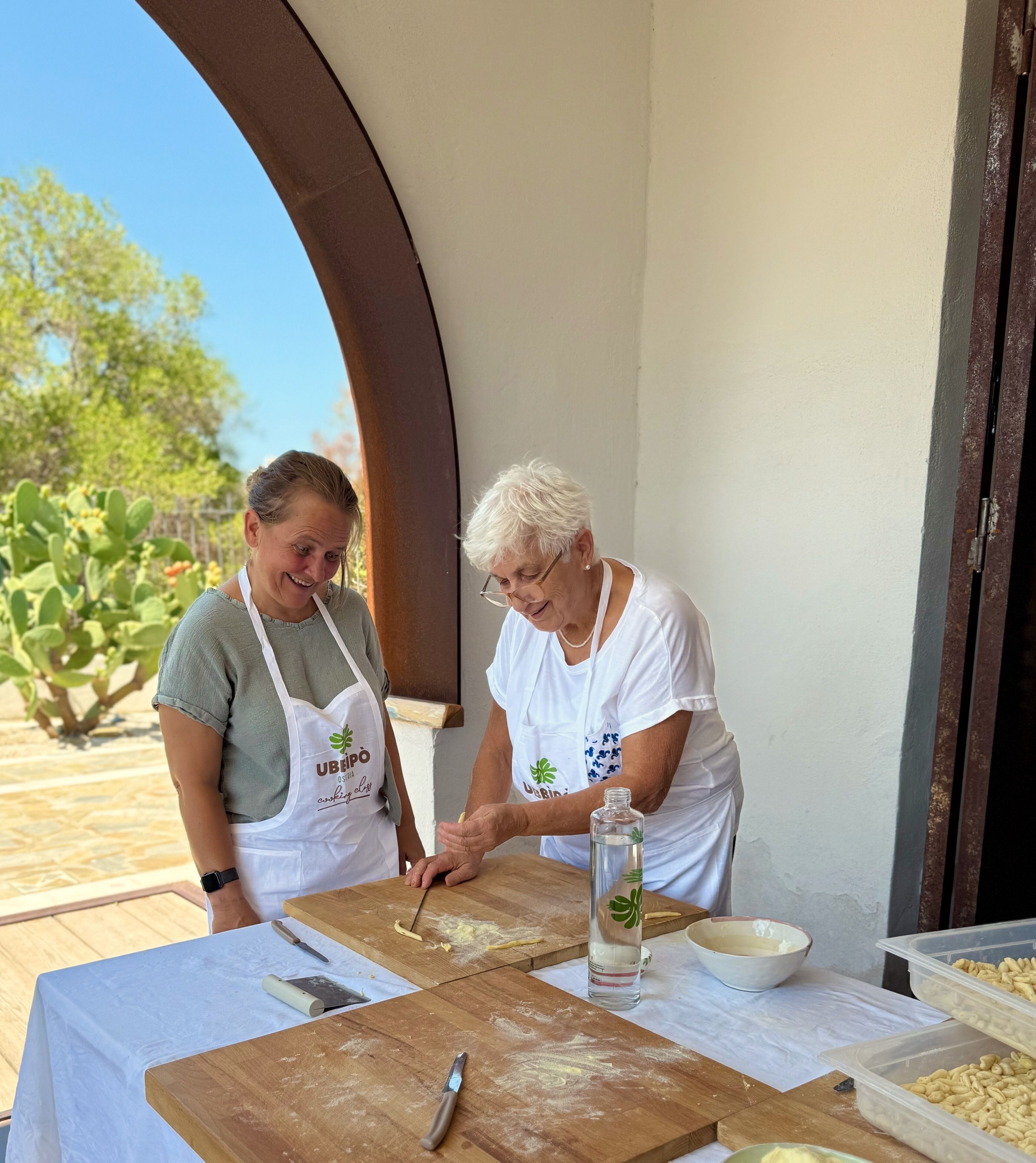 Two people preparing food outdoors with a table, a knife, and a bottle.