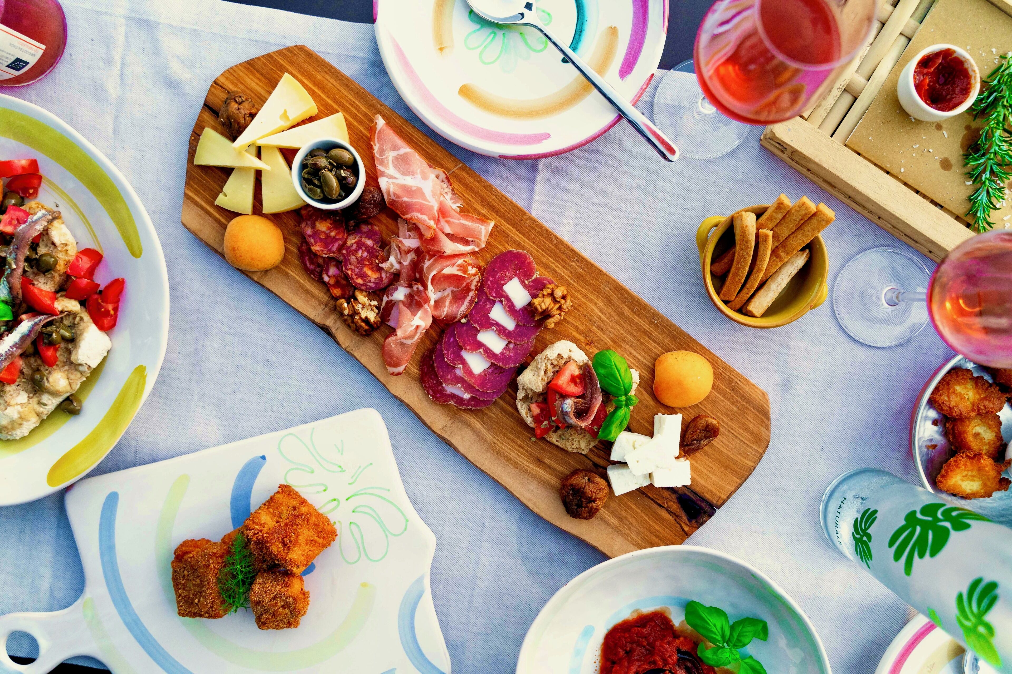 A variety of food items on a wooden board, including cheese, cured meats, and bread, with wine glasses and a bowl of olives in the background.