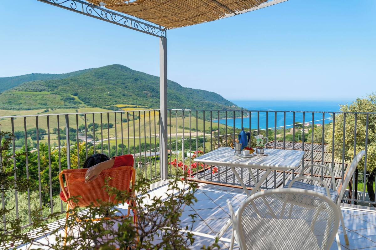 Une terrasse avec une table et des chaises, offrant une vue sur un paysage côtier avec des collines et la mer.