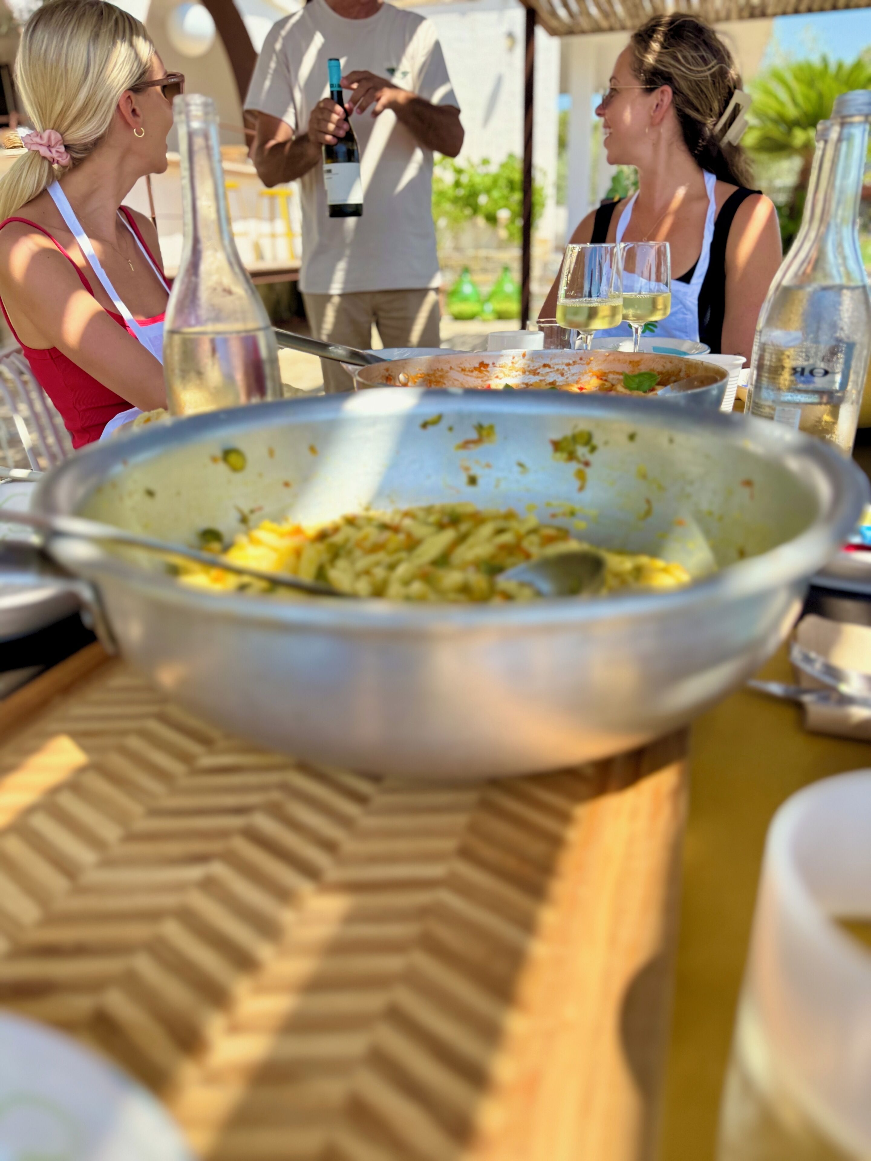 A dining table with a large bowl of pasta, wine bottles, and glasses.
