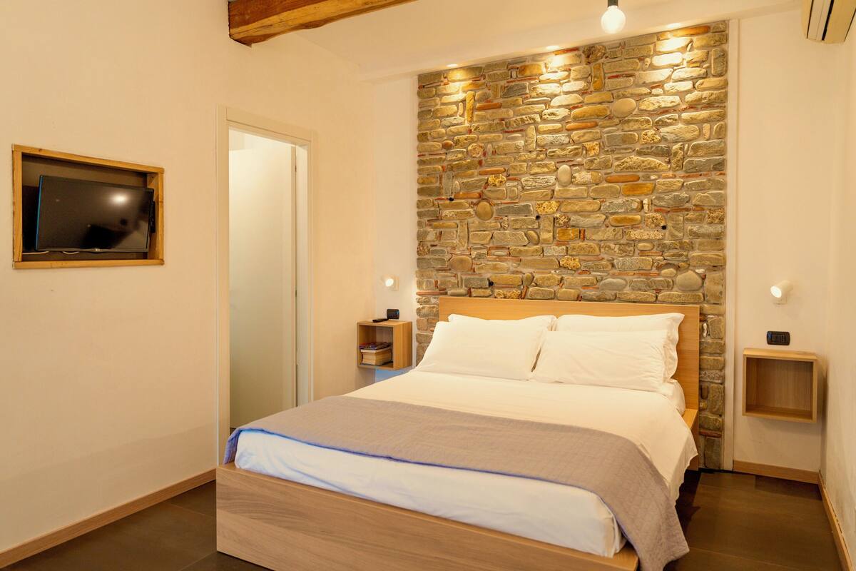 Une chambre avec un mur en pierre, un cadre de lit en bois, un téléviseur fixé au mur et des tables de chevet.