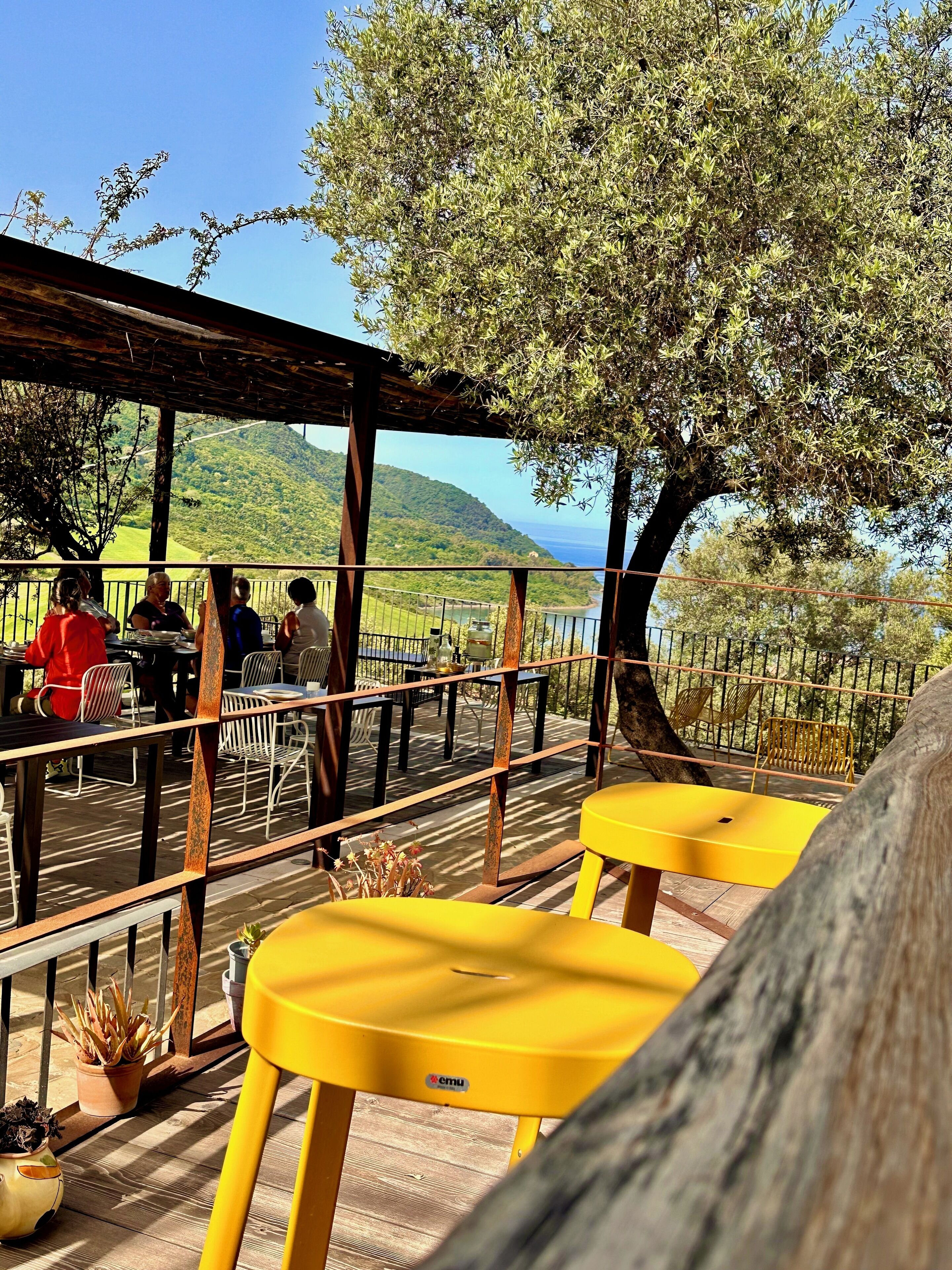 An outdoor seating area with yellow tables and chairs, overlooking a scenic landscape with hills and a body of water.