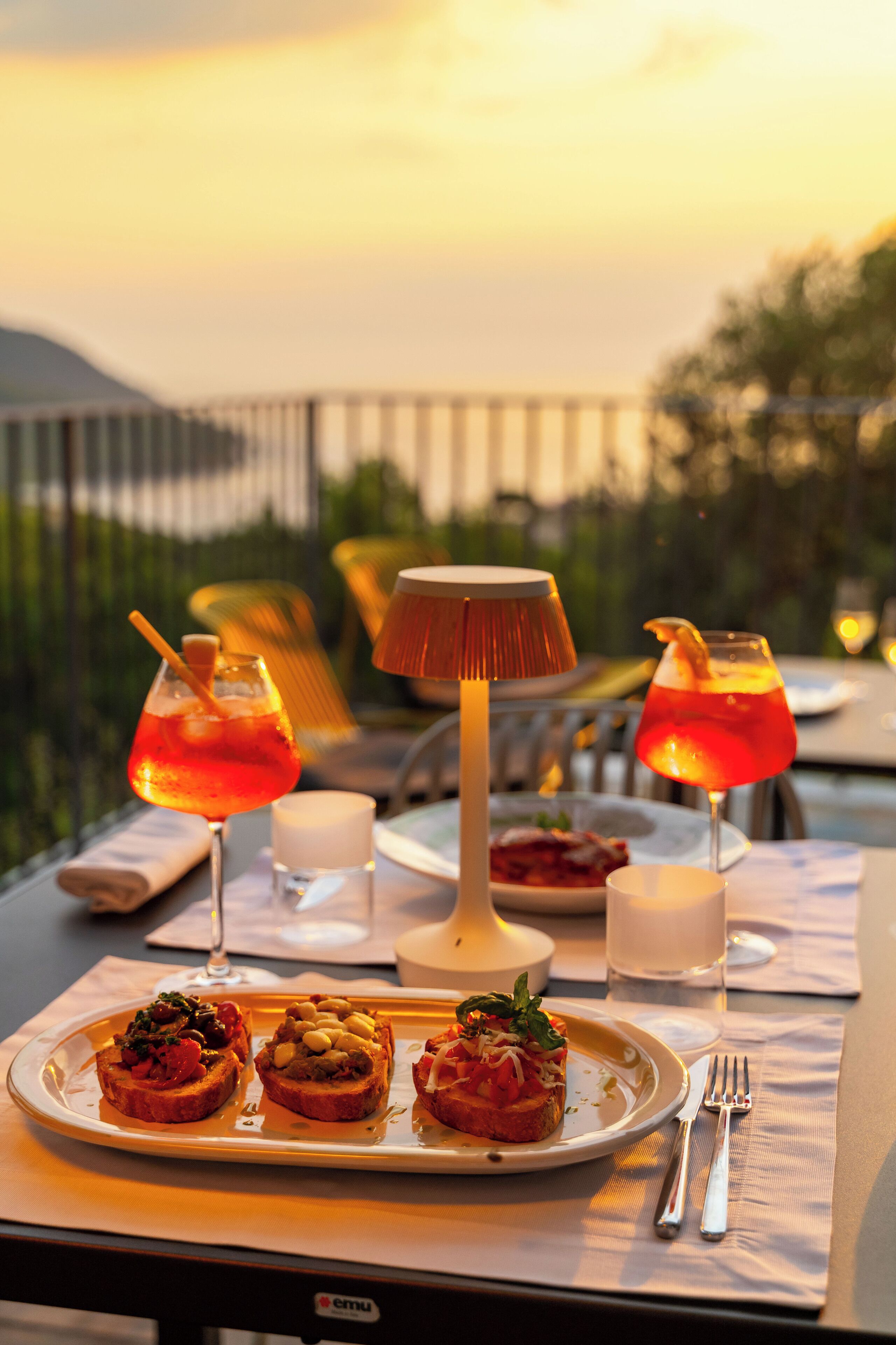 Una mesa servida con bruschetta, dos bebidas, una vela y una lámpara.