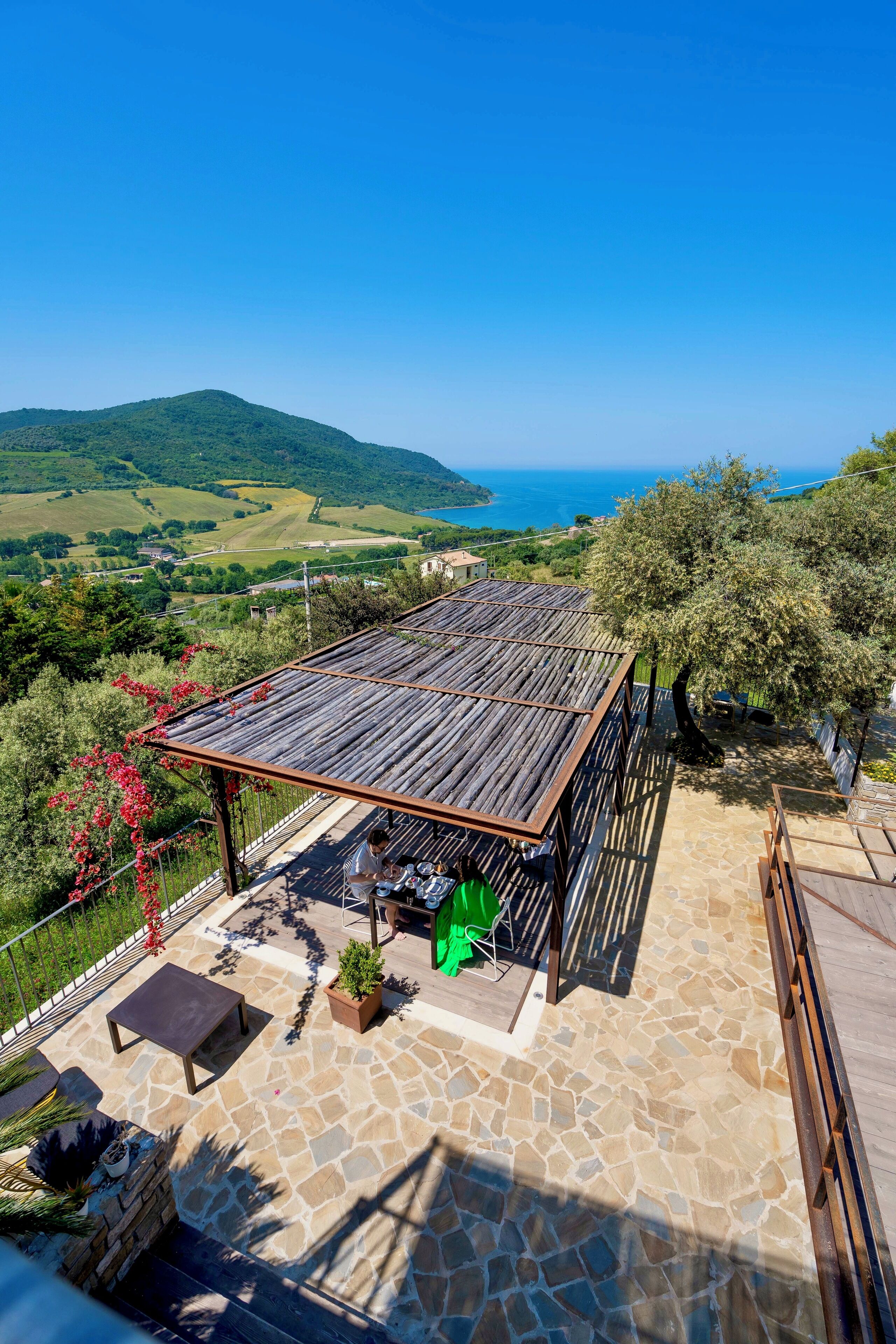 A stone patio with a wooden pergola, outdoor seating, and a view of the sea and hills.