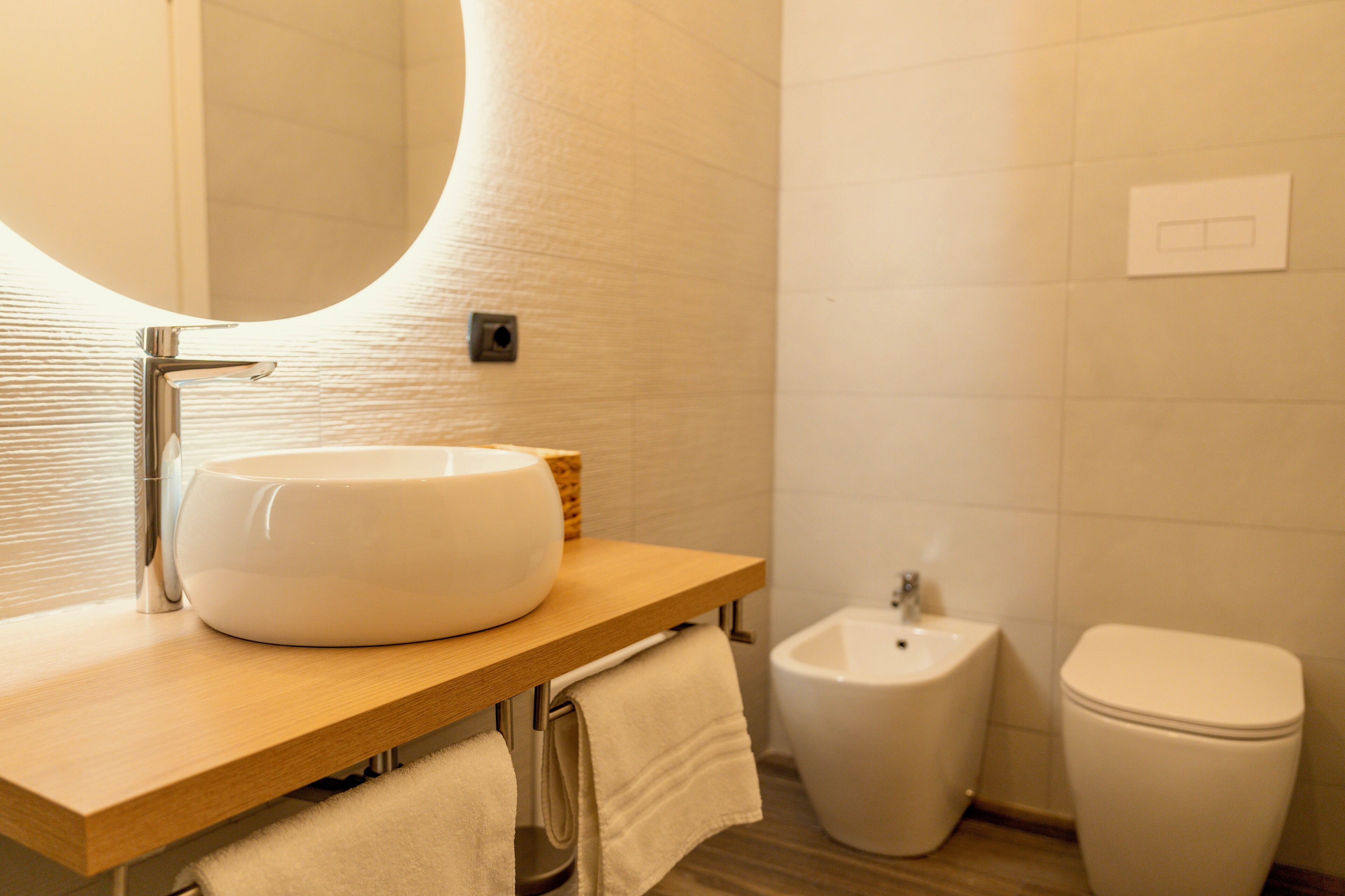 A modern bathroom with a round mirror, a white sink, a bidet, and a toilet.