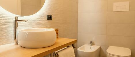 A modern bathroom with a round mirror, a white sink, a bidet, and a toilet.