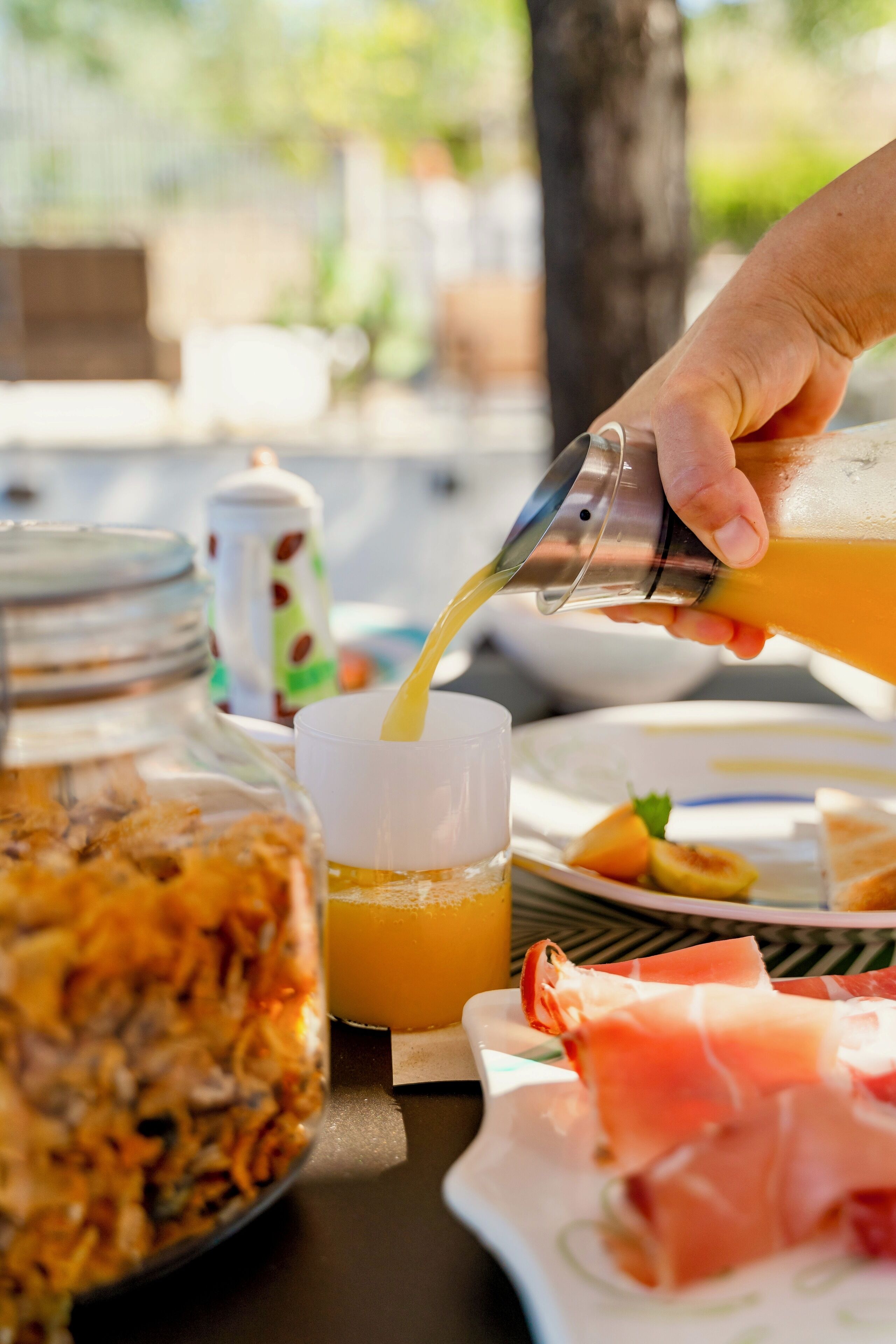 Une table mise pour le petit-déjeuner, avec une main versant du jus d’orange dans une tasse, une assiette de fruits coupés et un bocal de granola.