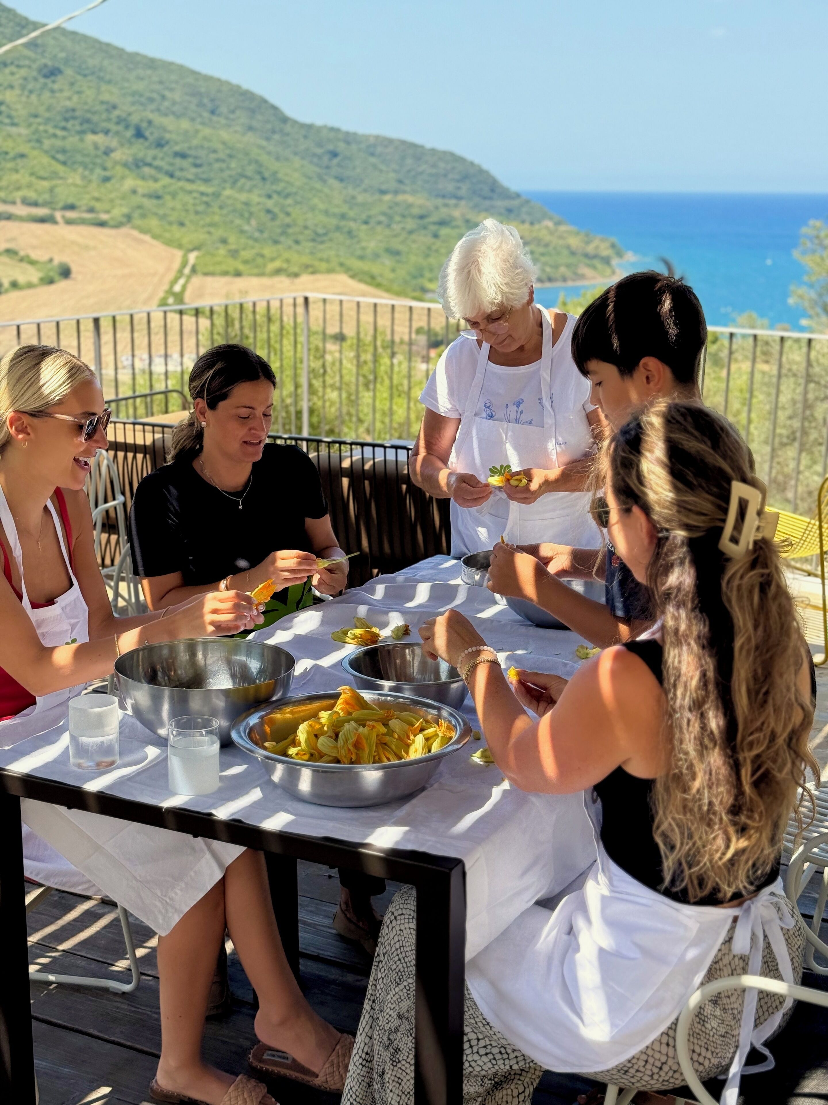 A group of people dining outdoors with a table set for a meal, surrounded by a scenic view of hills and the sea.