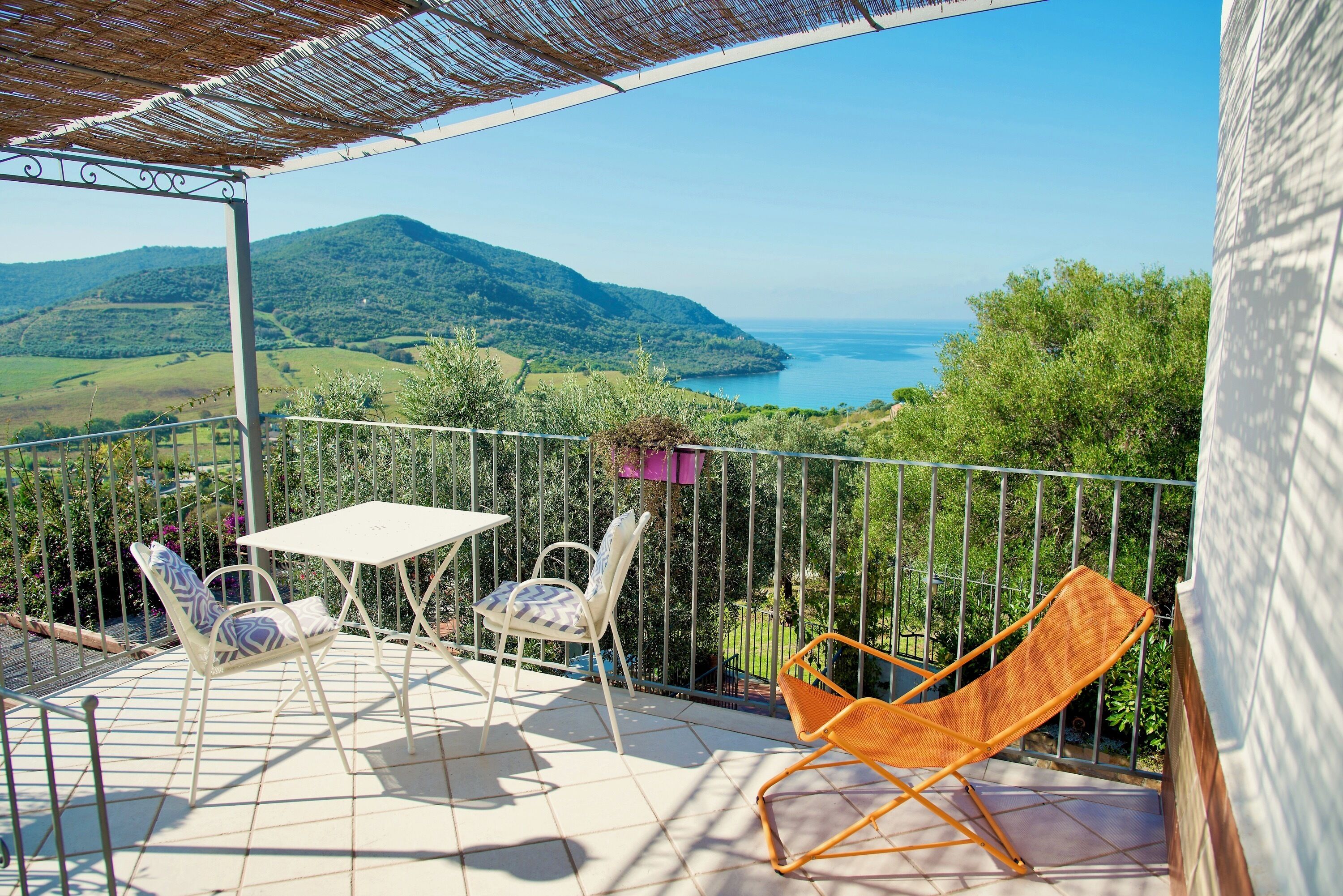 A balcony with a table and chairs, a hanging plant, and a view of the sea and hills.