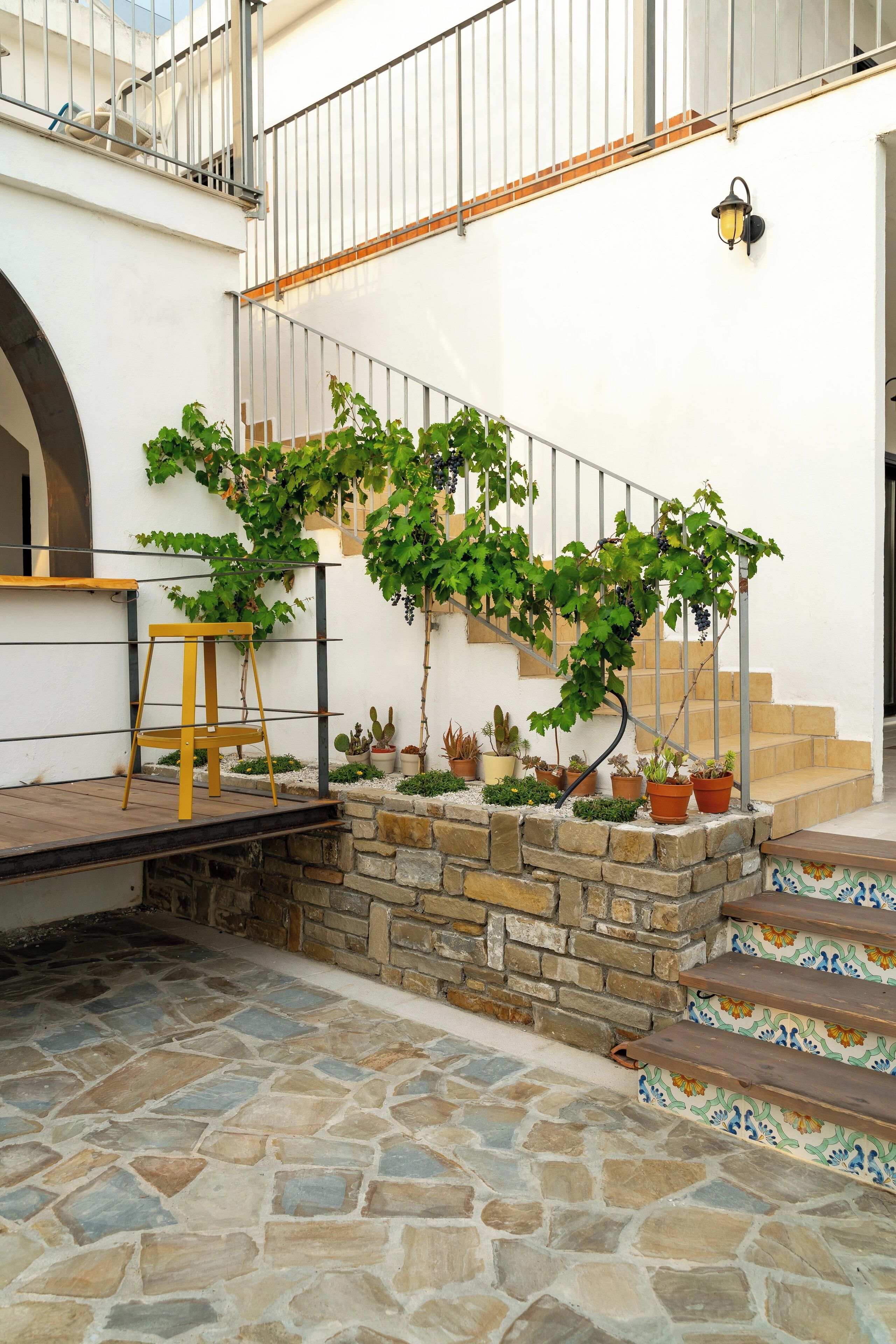 A stone staircase leading up to a white wall with a metal railing, surrounded by potted plants and a wooden stool.