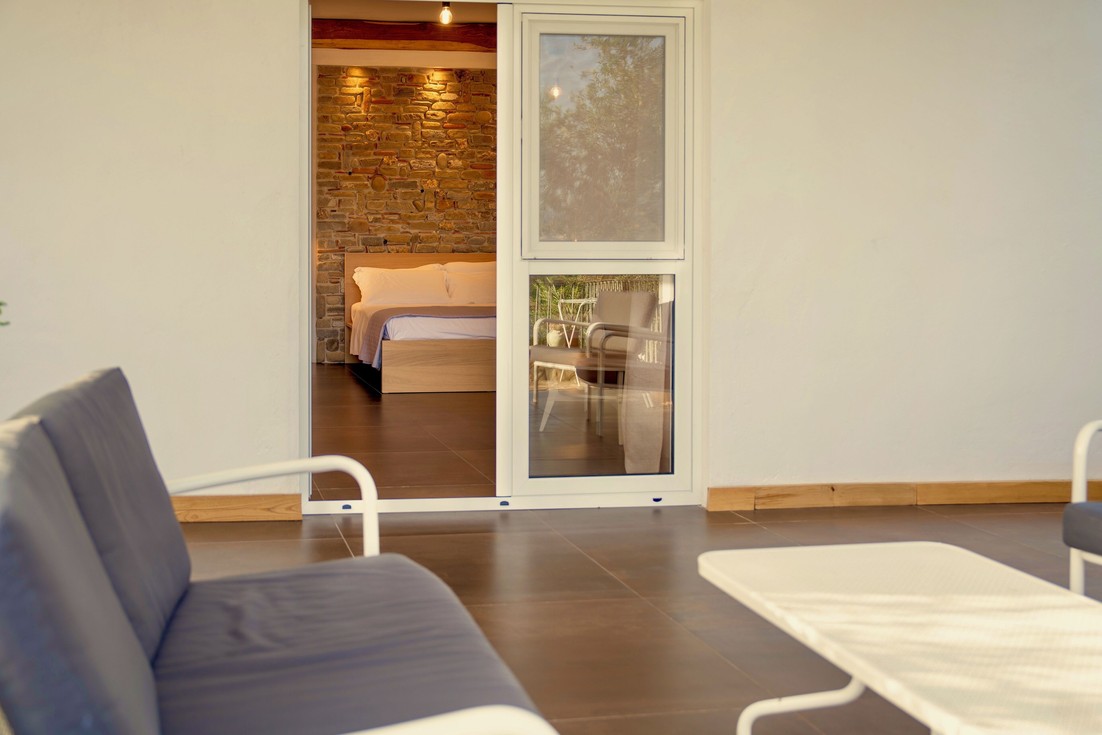 Ein modernes Zimmer mit grauem Sofa, einem kleinen weißen Tisch und einer Glastür, die auf einen Balkon mit Stuhl und Tisch führt.