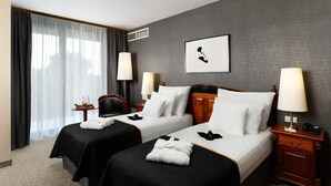A hotel room with two beds, a large window with curtains, a framed picture on the wall, and a nightstand with a lamp.