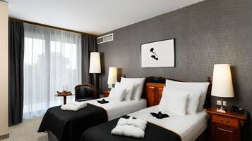 A hotel room with two beds, a large window with curtains, a framed picture on the wall, and a nightstand with a lamp.