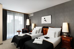 A hotel room with two beds, a large window with curtains, a framed picture on the wall, and a nightstand with a lamp.