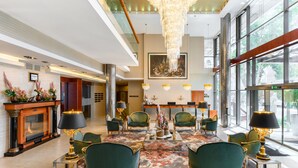 A spacious lobby with a large chandelier, green upholstered chairs, a fireplace, and large windows offering a view of the outdoors.