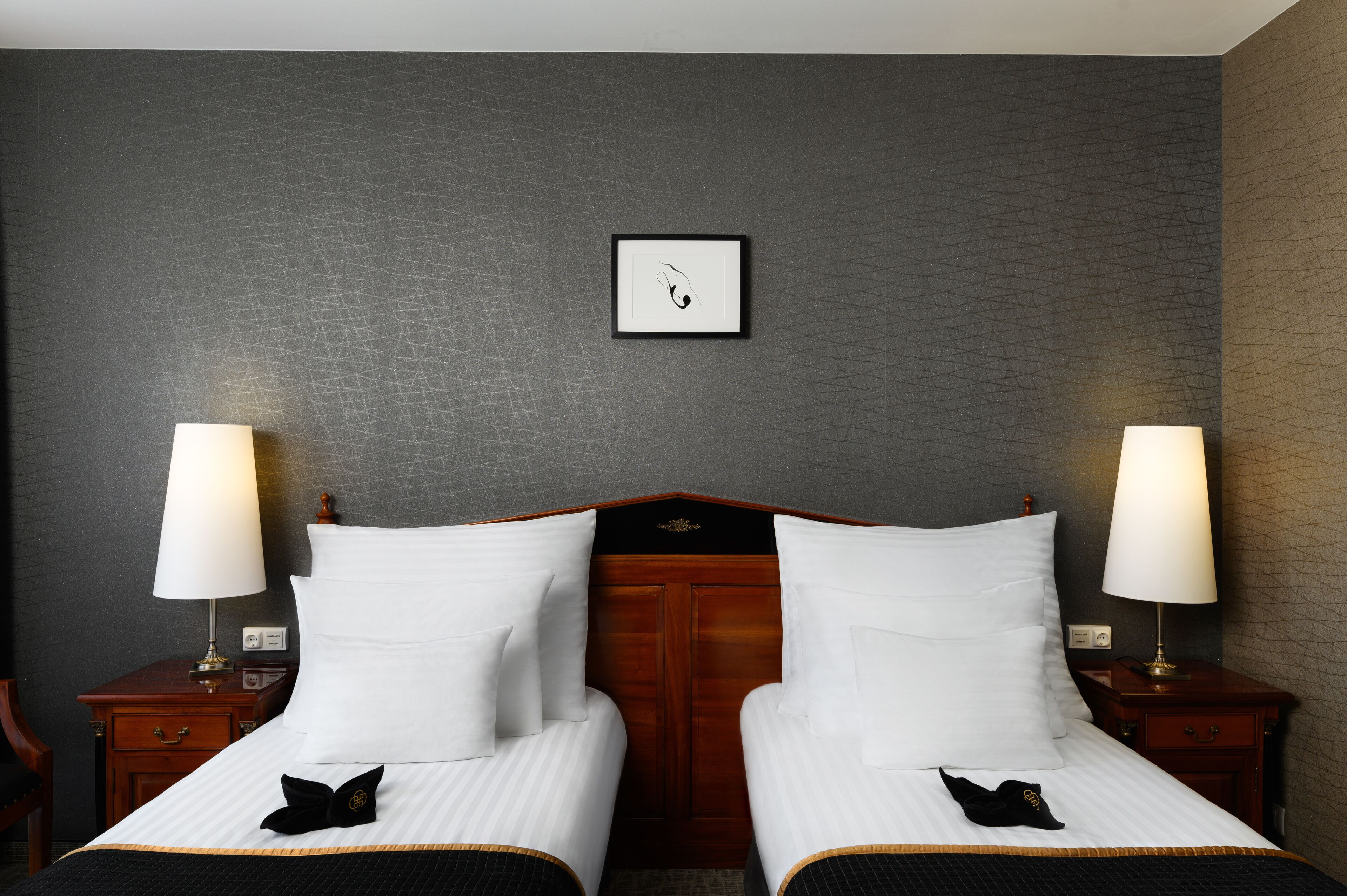 A hotel room with two beds, each with white linens and black decorative pillows. There are bedside lamps on wooden nightstands.