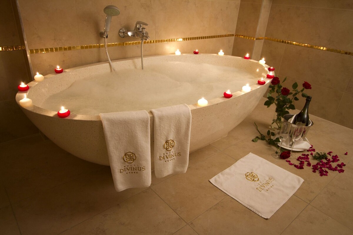 Une grande baignoire ovale entourée de bougies, deux serviettes portant la marque « Hotel Divinus », et une bouteille et des verres posés à proximité.
