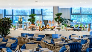 A modern cafe with blue and beige upholstered chairs, a central buffet with various food items, and large windows offering a view of the outdoors.