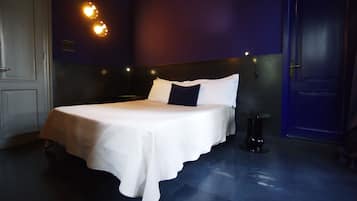 Un lit bien fait, recouvert d’une couvre-lit blanche et agrémenté d’oreillers, une petite table ronde placée à côté, et un luminaire fixé au mur.