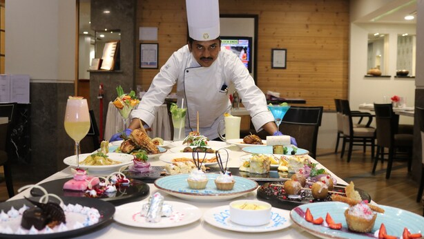 Un chef, vêtu d'un uniforme blanc et coiffé d'un haut-de-forme, prépare des plats sur une table où sont disposés divers mets et boissons.