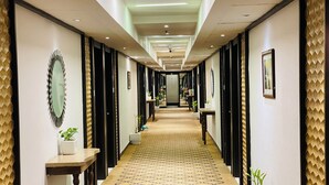 Un couloir d’hôtel avec un tapis à motifs, des plantes en pot et des tableaux encadrés accrochés aux murs.