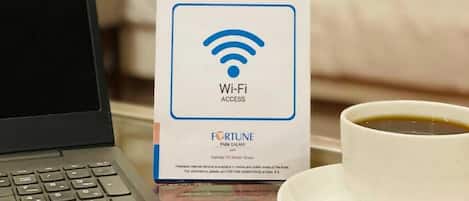 A laptop on a table with a Wi-Fi access sign, a cup of coffee, and a bed with pillows in the background.