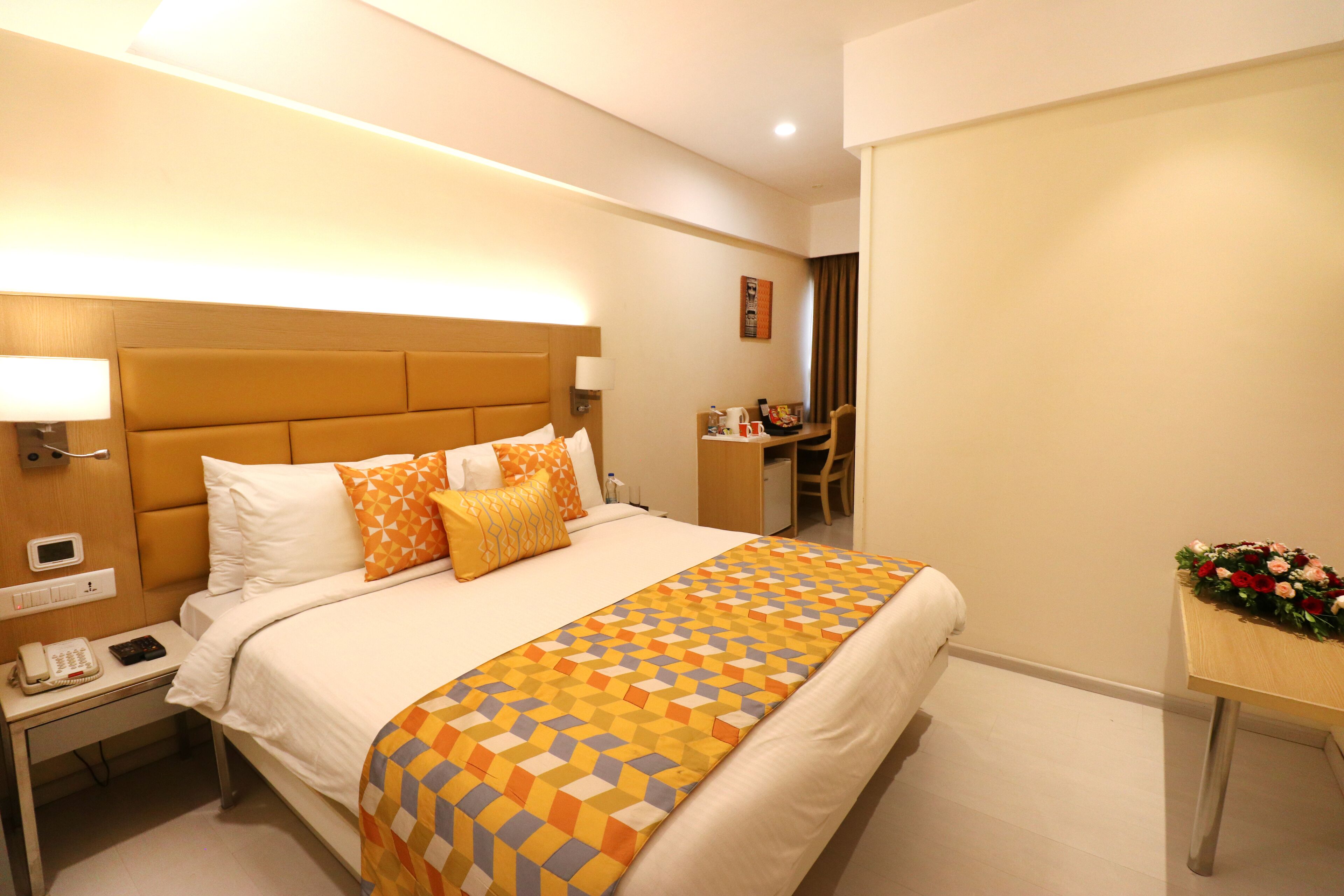 A hotel room with a large bed, a bedside table with a phone, a desk with a chair, and a small table with flowers.