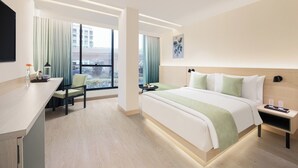 Une chambre d’hôtel moderne avec un grand lit, un bureau et une chaise. Elle dispose de grandes fenêtres avec des rideaux, d’un téléviseur à écran plat et d’une petite table avec une lampe.