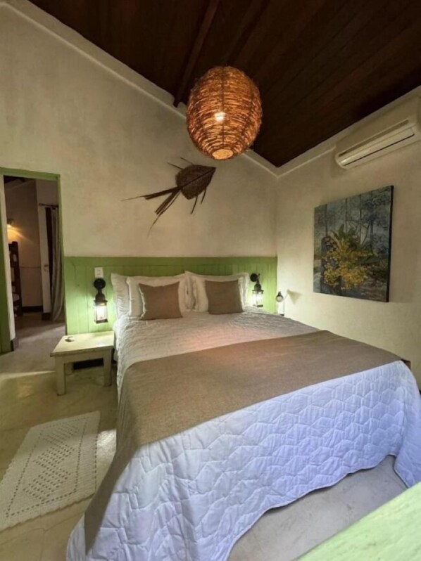 A bedroom with a large bed, green headboard, two bedside lamps, a woven ceiling light, a wall-mounted air conditioner, and a painting.
