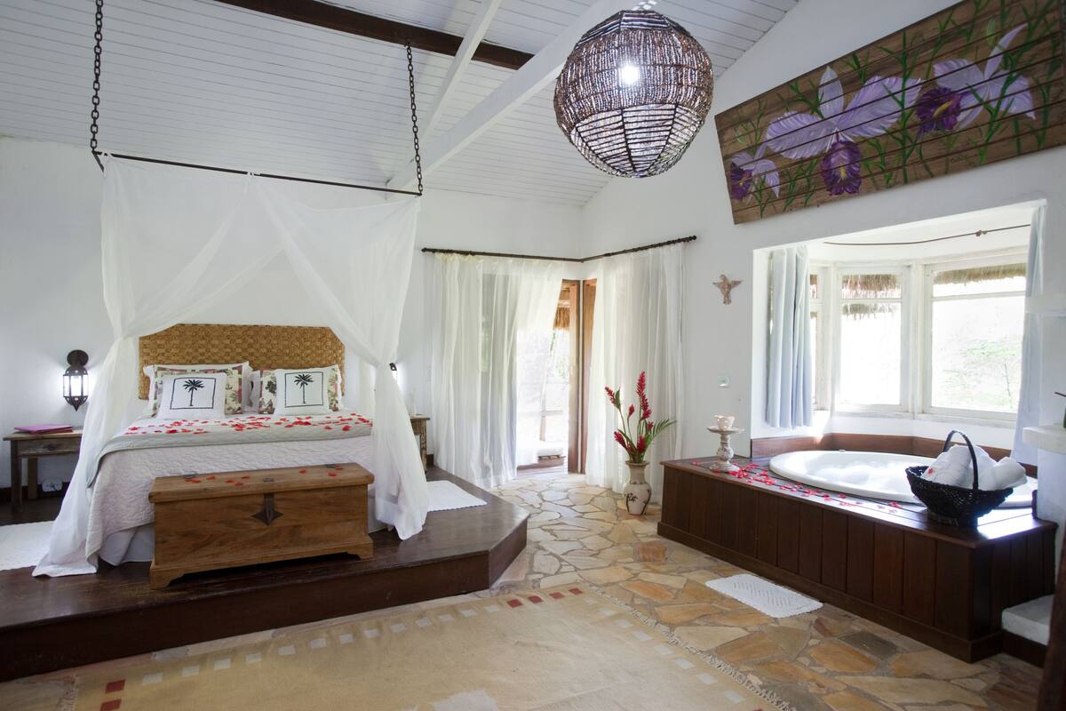 A bedroom with a bed, a stone floor, a wooden bench, a bathtub, and a hanging light fixture.