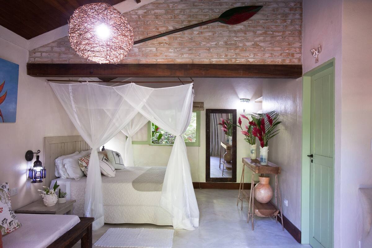 A bedroom with a bed under a white canopy, a bedside table with a lamp, a mirror, and a green door.